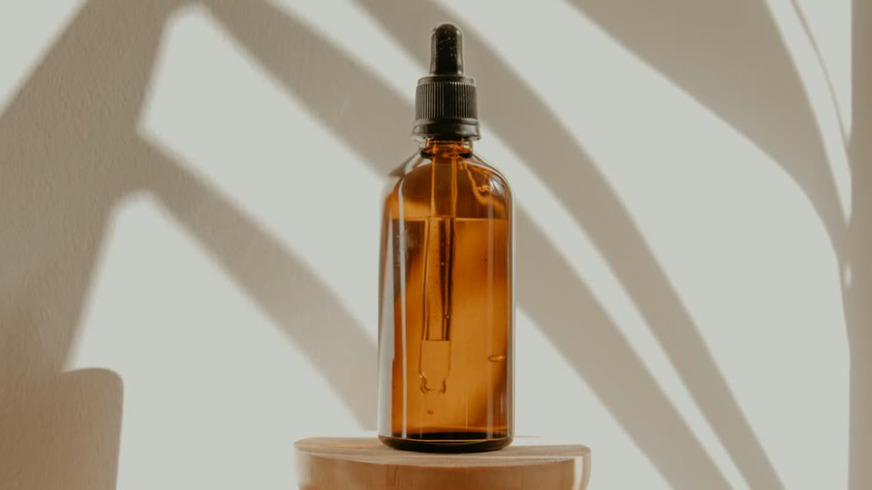 Frasco âmbar com conta-gotas sobre um pedestal de madeira, com sombras de folhas na parede.
